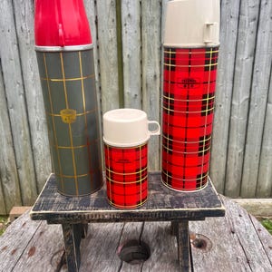 May include: Three vintage Thermos brand vacuum flasks in a variety of sizes. One is olive green with gold grid lines and a red cap. The other two are red plaid with cream-colored lids. The flasks are displayed on a small, black, wooden table.