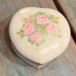 May include: Heart-shaped trinket box with a cream-colored finish. The lid features a hand-painted bouquet of pink roses with green leaves. The box has a gold trim and the name "E. Klein" is written on the bottom.