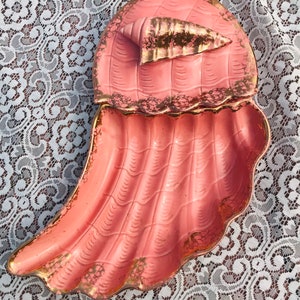 May include: A pink ceramic seashell dish with gold accents. The dish is divided into two sections, one for holding a small seashell and the other for holding other items.