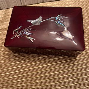 May include: A rectangular, burgundy-colored jewelry box with a glossy finish. The lid features a decorative inlay of a bird perched on a branch with iridescent accents. The box is set on a textured, brown surface.