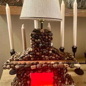 May include: A stone fireplace lamp with a white lampshade. The fireplace has a red light inside, simulating a fire. The lamp is on a wooden surface with candles on either side.