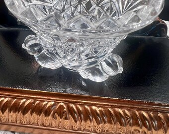 May include: A clear, cut-glass bowl with a scalloped rim and a starburst pattern. The bowl sits on a pedestal base with three decorative feet. The bowl is likely used for serving fruit or candy.