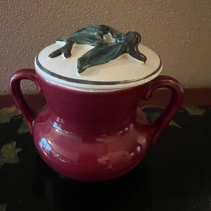 May include: A burgundy ceramic sugar bowl with a white lid and a green leaf design. The lid has a raised design of two leaves.