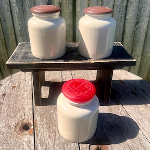 May include: Three ceramic jars with lids. Two jars have brown lids and are on a small black wooden table. The third jar has a red lid and is on a wooden surface. The jars are off-white.