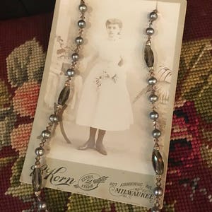 May include: A vintage photo of a young girl wearing a white dress, with a pearl and dark brown glass bead necklace draped over the photo. The photo is on a card with the text "Korn Extra Finish 907 Kinnickinnic Ave. Milwaukee."
