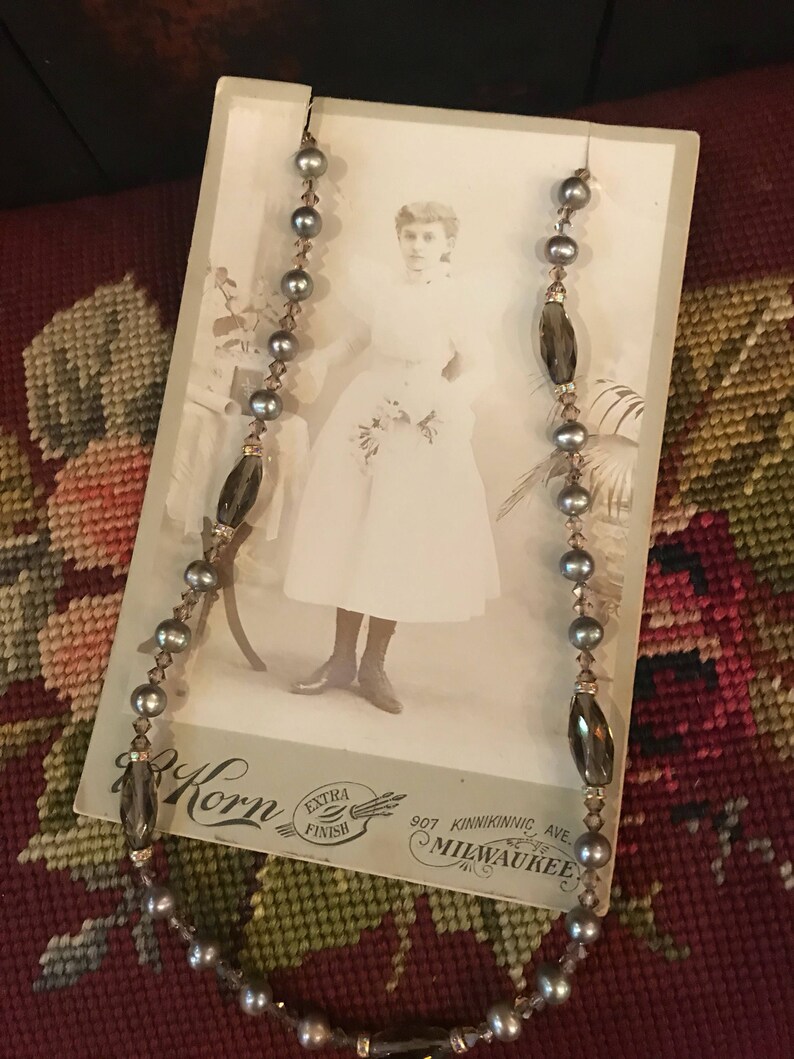 May include: A vintage photo of a young girl wearing a white dress, with a pearl and dark brown glass bead necklace draped over the photo. The photo is on a card with the text "Korn Extra Finish 907 Kinnickinnic Ave. Milwaukee."