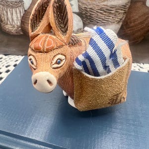 May include: A ceramic donkey figurine with large ears, blue eyes, and a tan body. A small pouch on its back holds a blue and white striped cloth. The figurine is on a blue surface.