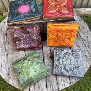 May include: An assortment of square, decorative tiles with embossed floral designs. The tiles are in various colors, including red, orange, green, purple, and gray. Some tiles have a glossy finish, while others appear matte.