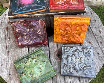 May include: An assortment of square, decorative tiles with embossed floral designs. The tiles are in various colors, including red, orange, green, purple, and gray. Some tiles have a glossy finish, while others appear matte.
