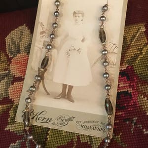 May include: A gray and brown beaded necklace with a vintage photo of a young girl in a white dress. The photo has the text "Korn Extra Finish 907 Kinnickinnic Ave Milwaukee" printed on the bottom.