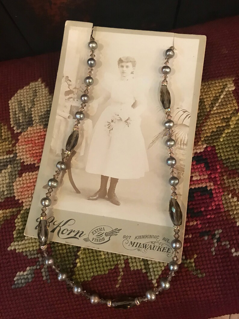 May include: A gray and brown beaded necklace with a vintage photo of a young girl in a white dress. The photo has the text "Korn Extra Finish 907 Kinnickinnic Ave Milwaukee" printed on the bottom.