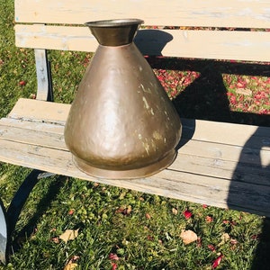 Puede incluir: Un jarrón grande de color cobre con textura martillada se encuentra sobre un banco de madera. El jarrón tiene una abertura ancha y acampanada y un fondo redondeado.