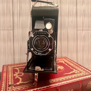 May include: A black folding camera with a leather exterior and a lens with the text "No. 1 KODON" and "T B 25 50 100". The camera is sitting on top of a red book with gold lettering that reads "Oliver Twist".