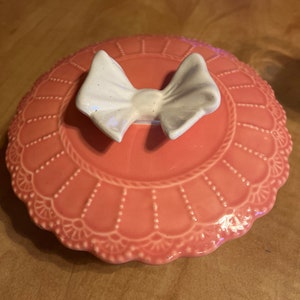 May include: A pink ceramic lid with a white bow knob. The lid has a scalloped edge and a delicate floral design.
