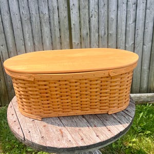 May include: A light brown, oval-shaped woven basket with a matching wooden lid. The basket is made of woven strips of wood and has a sturdy construction. It sits on a weathered wooden table, suggesting outdoor use. The basket is a storage container.