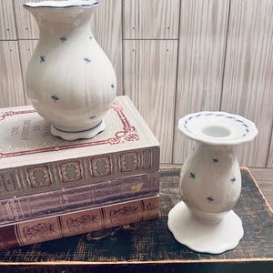 May include: Two white ceramic vases with blue floral patterns. The larger vase is taller and wider than the smaller vase. The smaller vase has a candle holder design. Both vases are sitting on a wooden table with a stack of books.