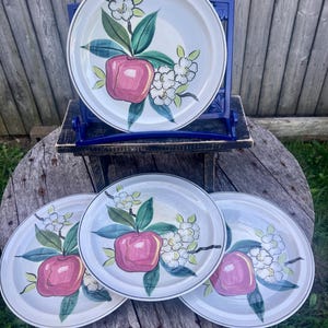 May include: Set of four ceramic plates with a hand-painted apple and floral design. Each plate features a pink apple, green leaves, and white flowers on a light blue background. The plates have a dark rim and are displayed on a wooden table.