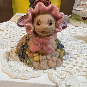 May include: A ceramic figurine of a smiling creature with a pink flower-shaped hat and a pink bow. The creature is sitting on a bed of green leaves and colorful berries.