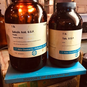 May include: Two amber glass jars with black lids and white labels. The labels read "1 lb. Salicylic Acid, U.S.P. Powder Tested for Mercury" and "1 lb. Talc, U.S.P. Powder". Both jars are from J.T. Baker Chemical Co., Phillipsburg, N.J. The jars are sitting on a stack of books with the title "HISTORY OF CEDARBURG".