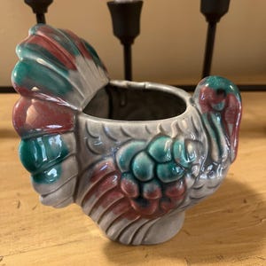 May include: A ceramic turkey-shaped planter with a gray base, teal and burgundy accents. The planter has a detailed feather design and a hollow interior. The turkey is positioned on a wooden surface.
