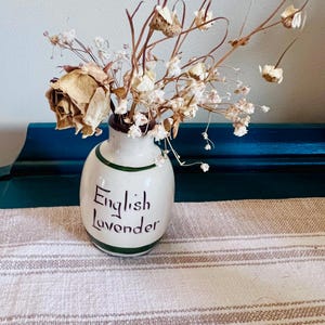 May include: A small, cream-colored ceramic jar with a narrow neck and dark green horizontal bands. The text "English Lavender" is written in black script on the front. It contains a dried floral arrangement with a beige rose and white baby's breath.
