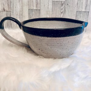 May include: A speckled ceramic mixing bowl with a handle and a dark blue rim. The bowl has a spout for easy pouring. The handle is a light gray color. The bowl is sitting on a white, fluffy surface.