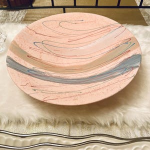 May include: A large, pink serving bowl with a speckled surface and a swirling design of blue, red, and green lines. The bowl is sitting on a white fluffy surface.