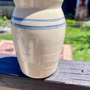 May include: A beige ceramic vase with two horizontal blue stripes. The vase has a slightly flared rim and a smooth, glossy finish. The background is blurred, showing a wooden surface and greenery.