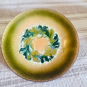 May include: A round, decorative plate with a unique design. The plate features a central peach-colored area surrounded by a swirling pattern of green and white. The outer rim is a darker shade of green, creating a striking contrast.
