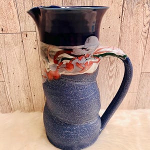 May include: A handcrafted ceramic pitcher with a dark blue glaze. The pitcher features a unique design with a textured body and a curved handle. The upper portion has a decorative pattern with red, white, and green accents. A functional and decorative piece.