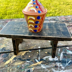 May include: A hand-painted ceramic jar with a lid, featuring a colorful design of red, blue, yellow, and white accents. The jar is sitting on a black wooden stool.