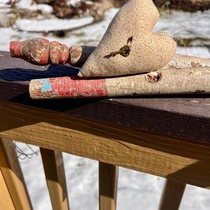 May include: A heart-shaped, beige fabric decoration with a key attached, resting on weathered wooden spindles. The spindles have red and blue paint. The background shows snow and a wooden fence.