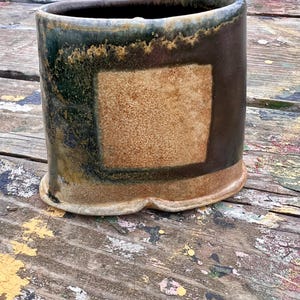 May include: A handcrafted ceramic vase with a rectangular design. The vase features a dark brown exterior with a lighter brown square in the center. The top rim has a dark green and brown glaze. The base has a unique scalloped design.
