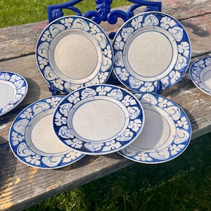May include: Set of six blue and white ceramic plates with a floral design. The plates are stacked on a wooden table.