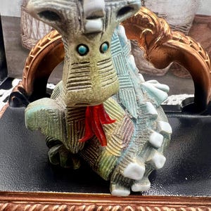 May include: A ceramic dragon figurine with a green, blue, and white patterned body. The dragon has a red tongue and blue eyes. The figurine is sitting on a black base with a gold trim.