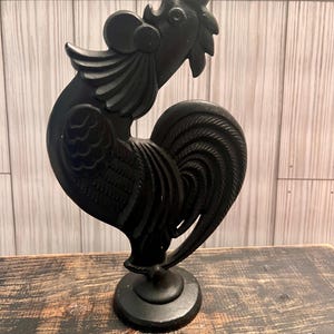 May include: A black cast iron rooster figurine with detailed feathers and a raised tail. The rooster is standing on a round base.