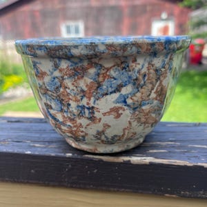 May include: A ceramic mixing bowl featuring a speckled, mottled pattern in shades of blue, brown, and cream. The bowl has a rounded, deep shape with a slightly flared rim and a small, raised circular base, showcasing a textured, multi-colored finish.