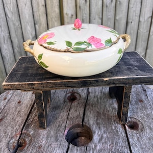May include: A white ceramic serving bowl with a lid, adorned with pink rose floral patterns and green leaves. The lid features a pink rose bud handle. The bowl has gold-colored handles and sits on a small, black wooden stand.