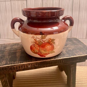 May include: A vintage ceramic crock with a brown rim and handles, and a cream-colored body. The crock features a fruit design with apples and grapes. The crock is sitting on a small, dark wooden stool.