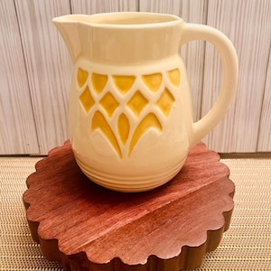 May include: A cream-colored ceramic pitcher with a yellow geometric design. The pitcher has a wide mouth and a handle. It is sitting on a wooden base.
