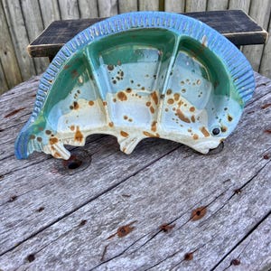 May include: A ceramic serving dish with three sections, featuring a blue, green, and white glaze with brown speckles. The dish is shaped like a fish and has a scalloped edge.