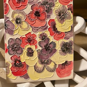 May include: A ceramic tile with a raised design of purple, yellow, and red pansies. The pansies are arranged in a repeating pattern.
