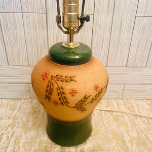 May include: A ceramic table lamp with a tan body, green base, and green top. The lamp features a floral design with red accents. The lamp has a brass-colored socket and a brown cord.