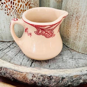 May include: A small, cream-colored ceramic pitcher with a red floral and scroll design around the rim. The pitcher has a curved handle and a spout for pouring. The pitcher is sitting on a rustic wooden surface.