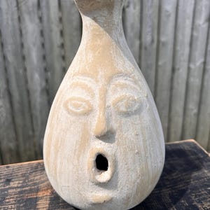 May include: A beige, handcrafted ceramic vase with a face design. The vase features carved eyes, a nose, and an open mouth. The vase has a narrow neck and a rounded body, and is likely used for decorative purposes.