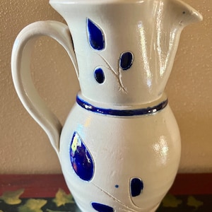 May include: A white ceramic pitcher with a blue glaze and a floral design. The pitcher has a handle and a spout.