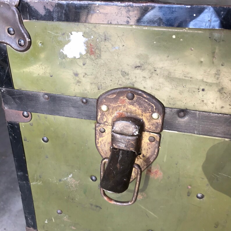 Puede incluir: Primer plano de un ba&uacute;l vintage de metal verde con un cierre y un asa de metal negro. El ba&uacute;l tiene un aspecto desgastado y envejecido con remaches visibles y un borde pintado de negro.