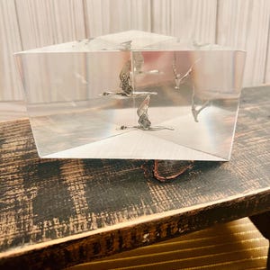 May include: A clear glass cube paperweight with three silver birds flying inside. The cube is sitting on a dark wooden surface.