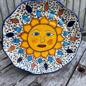 May include: A ceramic plate with a blue and white border and a yellow sun with a face in the center. The sun is surrounded by blue and brown teardrop shapes.