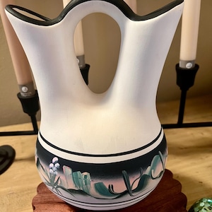 May include: White ceramic vase with a black rim and a handle. The vase has a southwestern design with green, pink, and black painted details.